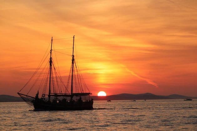 A silhouetted two-masted sailing ship carrying a WeRoad group trip sails on the ocean during a vibrant orange sunset.