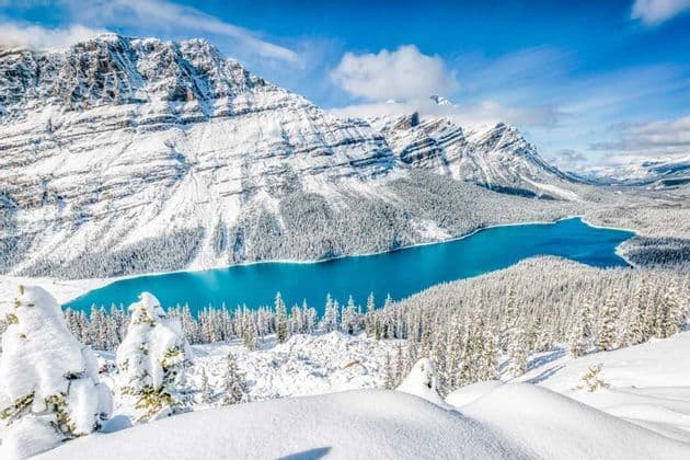 Un vivace lago turchese circondato da una vasta foresta innevata ai piedi di grandi montagne innevate.