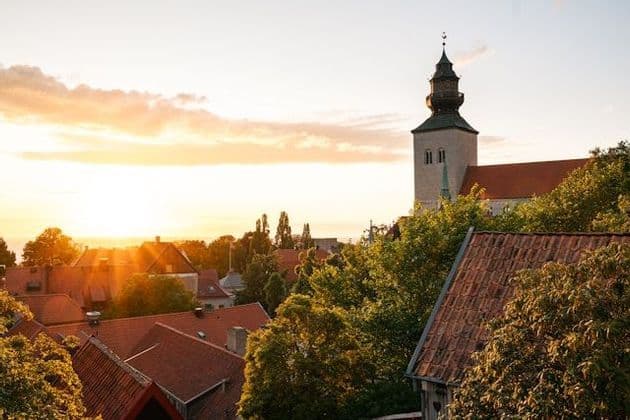Ein steinerner Kirchturm erhebt sich während eines goldenen Sonnenuntergangs über die roten Ziegeldächer und grünen Bäume einer Stadt.