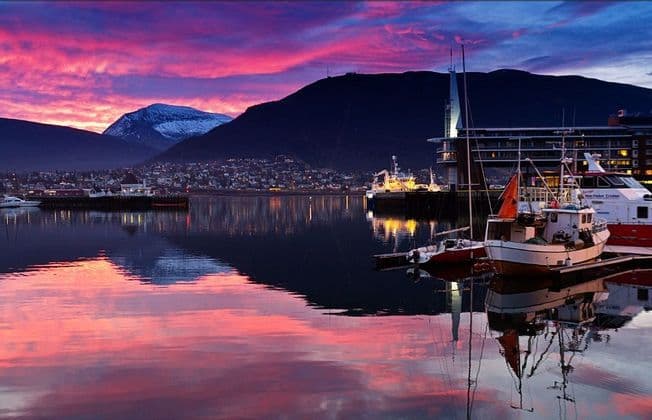 Un puerto con barcos amarrados refleja un vibrante cielo rosa y púrpura al atardecer, con un pueblo de montaña iluminado de fondo.