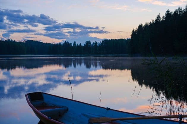 La prua di una piccola barca riposa sulla riva di un lago calmo al tramonto, con il cielo e una foresta riflessi sull'acqua.
