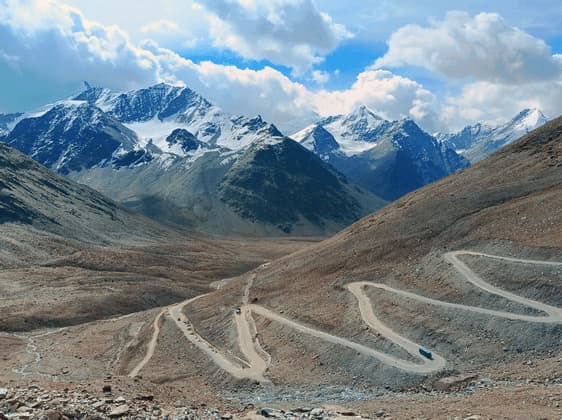 A winding dirt road snakes through a barren valley towards a range of snow-capped mountains under a cloudy sky.