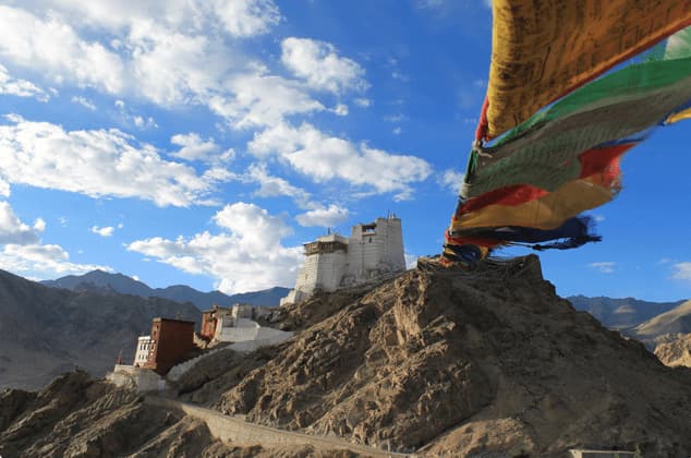 A white monastery sits atop a rocky mountain, with colorful prayer flags blowing in the wind in the foreground under a blue sky.