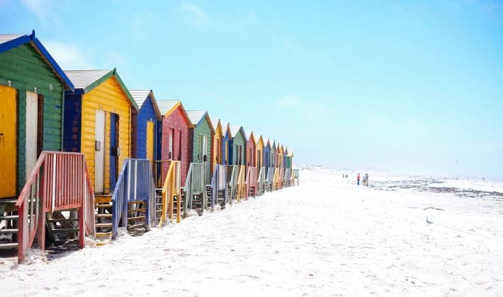 Une rangée de cabanes de plage en bois multicolores alignées sur une plage de sable blanc sous un ciel bleu clair.