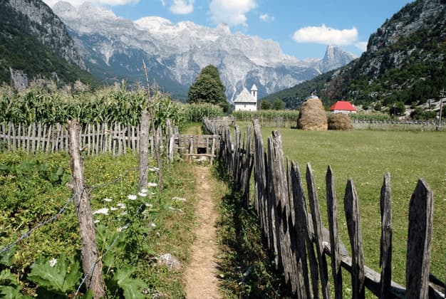 A dirt path runs alongside a rustic wooden fence through a green valley, leading towards a small church at the base of towering mountains.