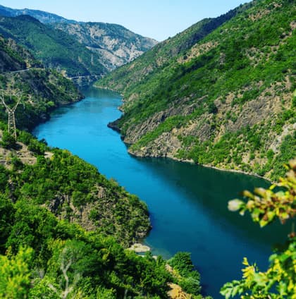An aerial view of a wide blue river winding through a valley of steep, green mountains under a clear sky.
