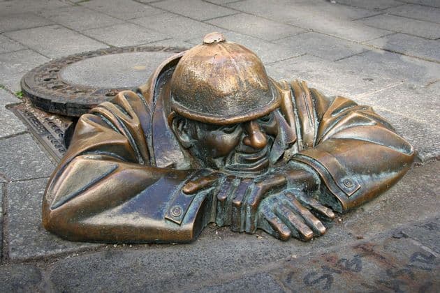 Una estatua de bronce de un hombre con sombrero emerge de una alcantarilla abierta en una acera pavimentada, apoyado en sus codos.