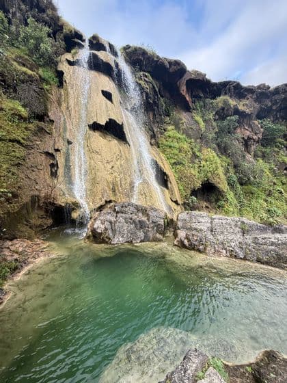 Una cascata scende lungo una parete rocciosa coperta di muschio in una piscina naturale limpida e verde sotto un cielo nuvoloso.