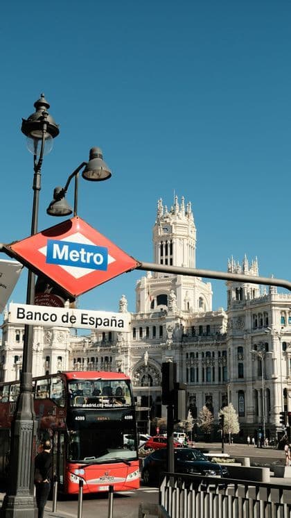 A red Metro sign for the Banco de España station, with a red tour bus and an ornate white building under a clear blue sky.