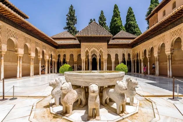 Una fuente de piedra con estatuas de leones se alza en un soleado patio de estilo morisco rodeado de columnatas arqueadas.