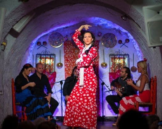 Una bailaora de flamenco con un vestido de lunares rojos actúa en un escenario dentro de una sala con arcos de piedra y músicos detrás.