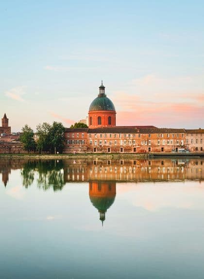 Un grande edificio in mattoni con una cupola verde si riflette perfettamente nell'acqua immobile di un fiume al tramonto.