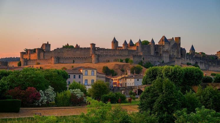 Un grand château de pierre avec de nombreuses tourelles se dresse sur une colline, illuminé par la lumière chaude du coucher de soleil au-dessus d'une petite ville aux arbres verdoyants.