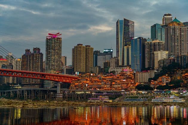 Uno skyline cittadino al crepuscolo con edifici tradizionali illuminati sulle colline e moderni grattacieli che si riflettono in un fiume vicino a un grande ponte rosso.