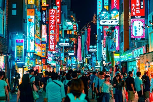 A crowd of people walks down a busy street at night, surrounded by tall buildings covered in bright, colorful Japanese neon signs.