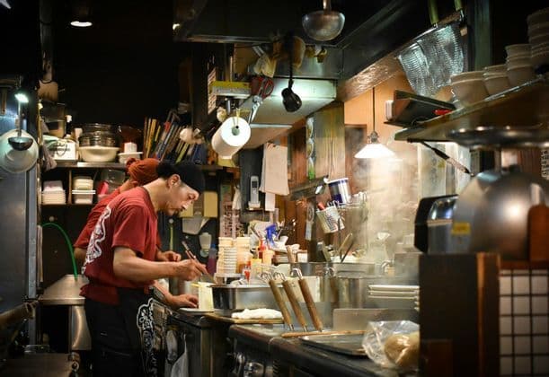 A chef in a red shirt and black bandana cooks with chopsticks in a busy, steamy restaurant kitchen filled with equipment.