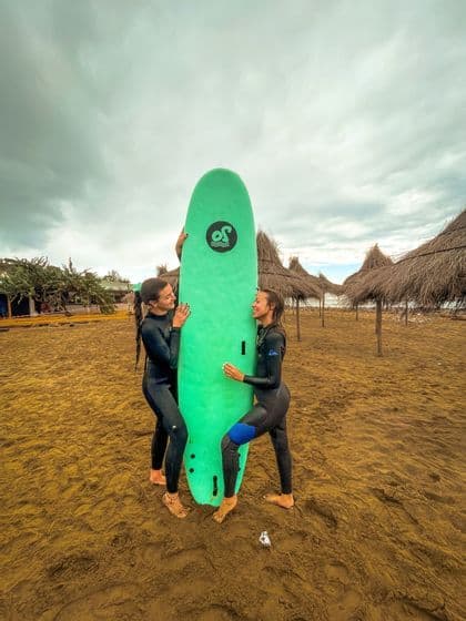Due donne di un viaggio di gruppo WeRoad, in mute, sorridono mentre tengono una tavola da surf verde in verticale su una spiaggia sabbiosa.