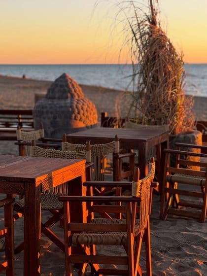 Tavoli in legno e sedie in vimini su una spiaggia di sabbia affacciata sull'oceano al tramonto, con una grande testa di Buddha in pietra lì vicino.