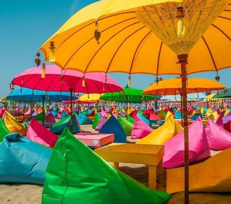Pouf colorati e grandi ombrelloni sono allineati su una spiaggia sabbiosa sotto un cielo limpido.