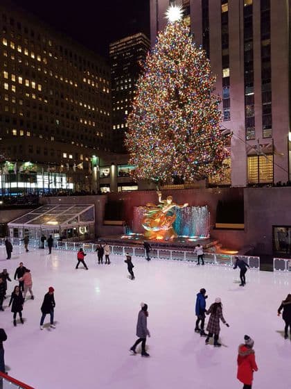 Des gens patinant sur glace la nuit sur une patinoire extérieure devant un grand sapin de Noël illuminé et des gratte-ciel de la ville.