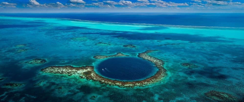 Una vista aerea di un grande, profondo buco blu marino circolare, circondato da un atollo di barriera corallina in un oceano turchese.
