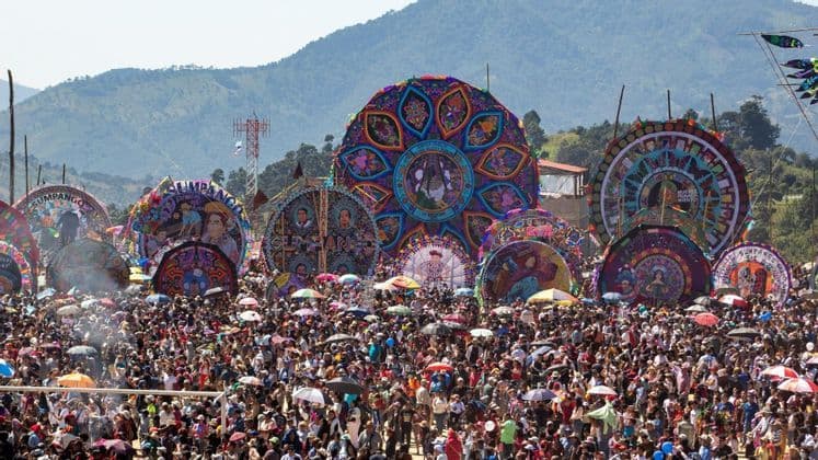 Eine riesige Menschenmenge versammelt sich auf einem Open-Air-Festival, um mehrere riesige, bunte, verzierte Drachen zu bestaunen, die vor einer Bergkette ausgestellt sind.