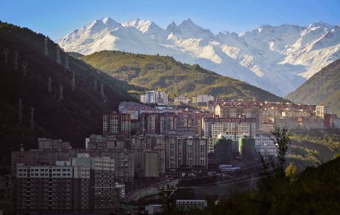 A city of apartment buildings built into a green mountain valley with a large, snow-capped mountain range in the background.