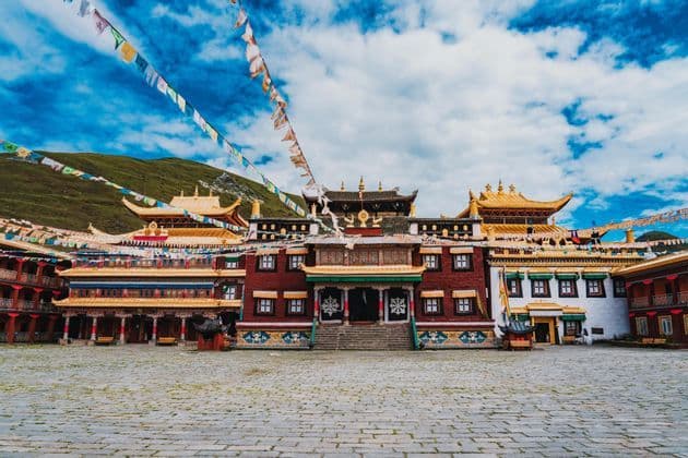 An ornate Buddhist monastery with golden roofs stands across a large stone courtyard, with prayer flags strung above under a blue sky.