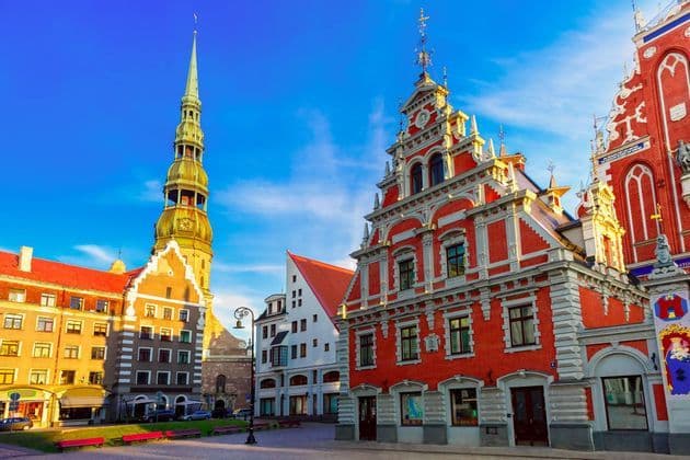 Des bâtiments ornés en briques rouges et un clocher d'église se dressent sur une place de ville sous un ciel bleu clair.