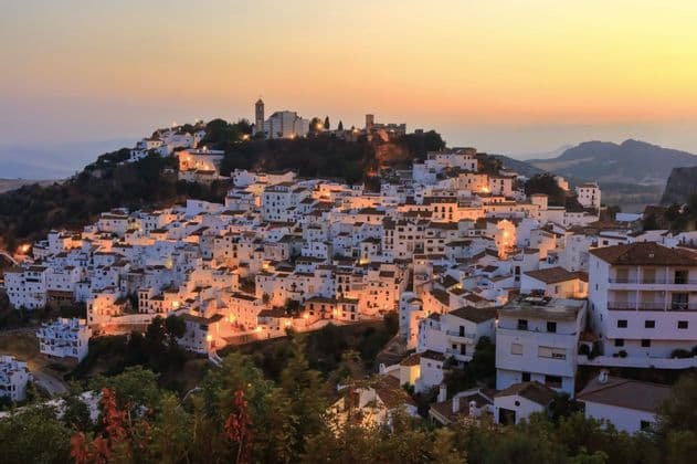 Un pueblo de casas de paredes blancas apiñadas en una ladera, con luces brillando al atardecer contra un cálido cielo anaranjado.