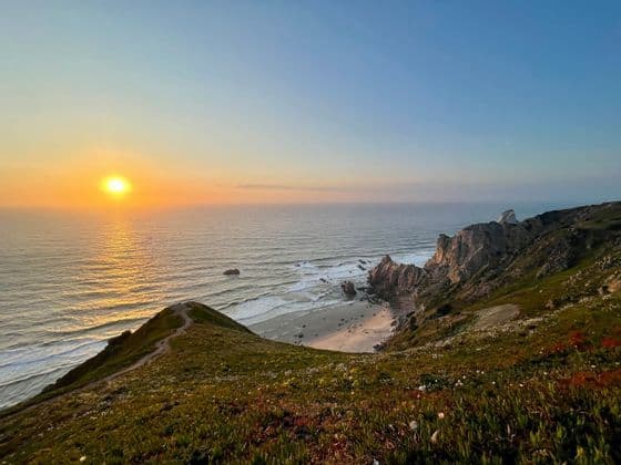 Il sole tramonta sull'oceano, visto da una scogliera erbosa con un sentiero che si affaccia su una spiaggia sabbiosa e una costa rocciosa.