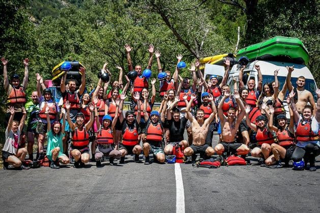 Un folto gruppo WeRoad in viaggio, con giubbotti di salvataggio e caschi, saluta per una foto di gruppo su una strada con gommoni sullo sfondo.