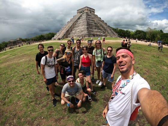 A WeRoad group trip takes a smiling selfie on a grassy field in front of a large ancient stone pyramid under a cloudy sky.