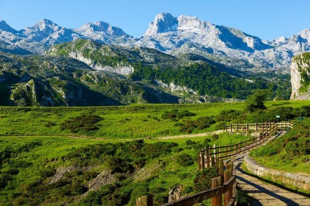 Un sendero de piedra con una valla de madera serpentea a través de un valle de montaña verde con picos rocosos al fondo bajo un cielo despejado.