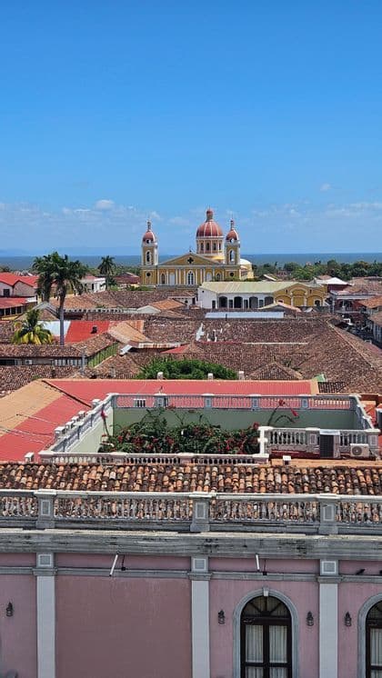 Eine gelbe Kathedrale mit roten Kuppeln erhebt sich über die terrakottafarbenen Dächer einer Stadt, von einem hohen Aussichtspunkt aus gesehen, mit Wasser in der Ferne.