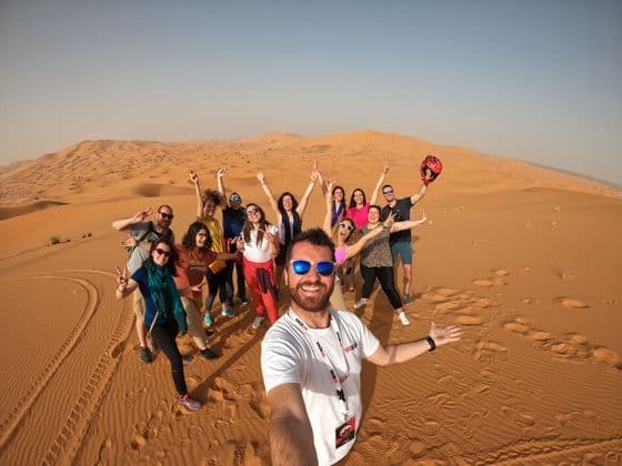 Un grupo de viaje de WeRoad sonríe y posa para una selfie en una gran duna de arena en el desierto bajo un cielo despejado.
