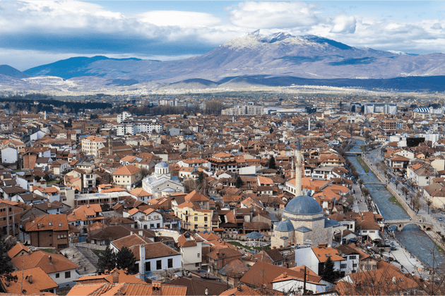 Una vista aerea di una città con edifici dai tetti in terracotta, una moschea e un fiume, con montagne innevate sullo sfondo.