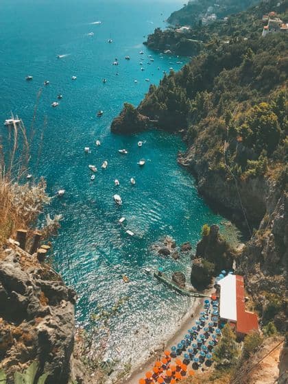 A high-angle view of a rocky coastline showing a cove with turquoise water, numerous boats, and a small beach with rows of colorful umbrellas.