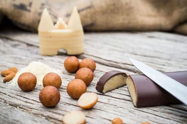 Tronchetto di marzapane al cioccolato affettato, accanto a palline di marzapane, mandorle tritate e uno stampo di pasta di mandorle, su un tavolo di legno.