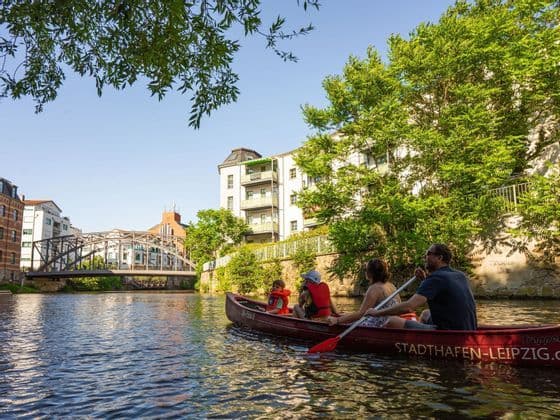 Eine vierköpfige Familie paddelt in einem roten Kanu auf einem Stadtkanal, gesäumt von Bäumen und Gebäuden.