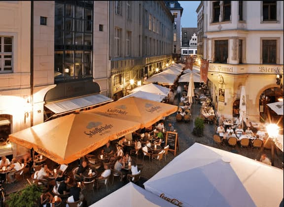 Eine Vogelperspektive auf eine belebte europäische Gasse, voller Menschen, die in Restaurants im Freien unter Sonnenschirmen in der Dämmerung essen.