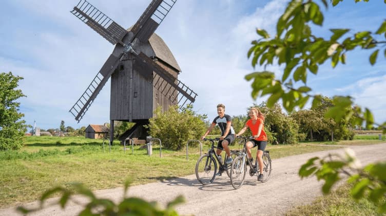 Ein Paar fährt auf einem Schotterweg auf dem Land Fahrrad, mit einer großen hölzernen Windmühle im Hintergrund.