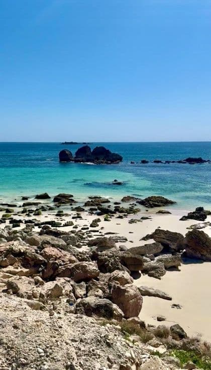 Una costa rocciosa incontra una spiaggia di sabbia bianca e acque turchesi sotto un cielo azzurro.