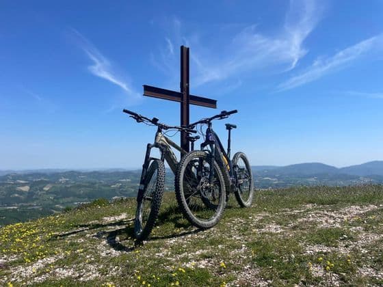 Due mountain bike parcheggiate su una cima erbosa accanto a una grande croce di metallo, con vista sulle dolci colline sotto un cielo azzurro.