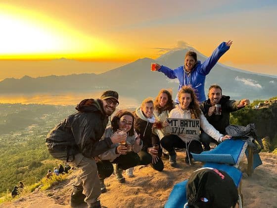 Un groupe WeRoad pose souriant pour une photo au sommet d'une montagne, des boissons à la main et un panneau indiquant le Mont Batur au lever du soleil.
