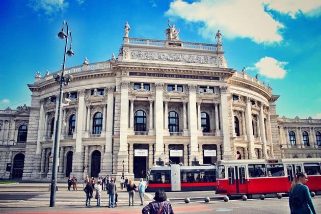 Un tramway rouge et blanc passe devant un grand et orné théâtre, sur une rue de ville animée de piétons.
