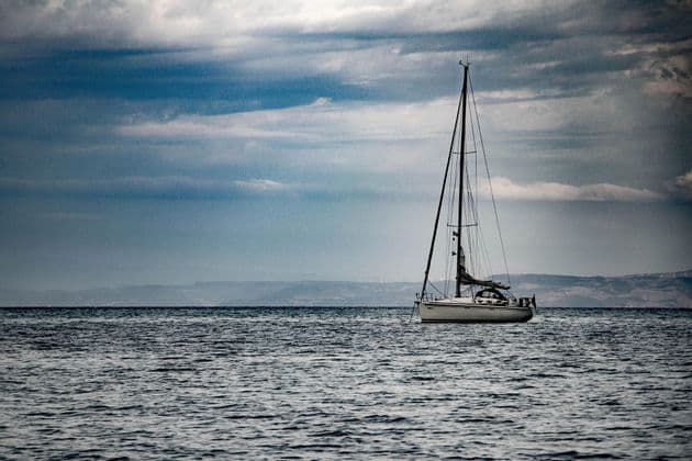 Una barca a vela bianca galleggia su acque agitate sotto un cielo nuvoloso e drammatico, con colline lontane all'orizzonte.