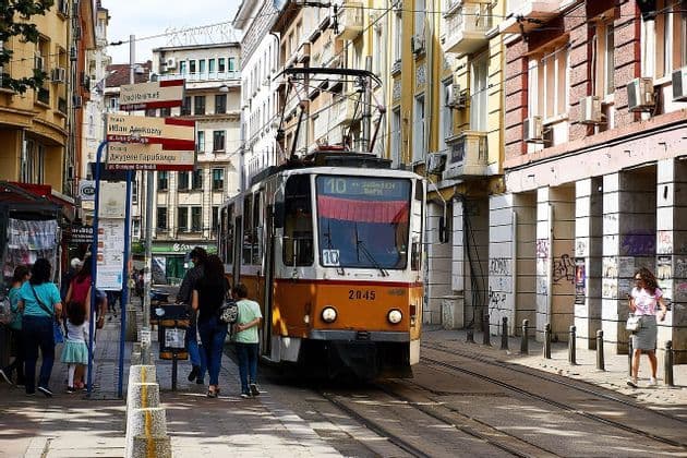 Eine gelb-weiße Straßenbahn fährt auf Schienen eine enge Stadtstraße entlang, mit Menschen, die auf dem Bürgersteig daneben gehen.