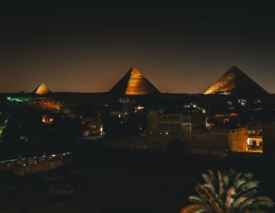 Vue nocturne de trois grandes pyramides illuminées de lumières dorées, s'élevant au-dessus d'un horizon urbain assombri.