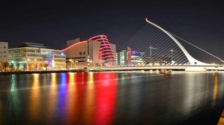 Eine moderne, beleuchtete Schrägseilbrücke überspannt nachts einen Fluss, wobei sich bunte Stadtlichter auf der Wasseroberfläche spiegeln.